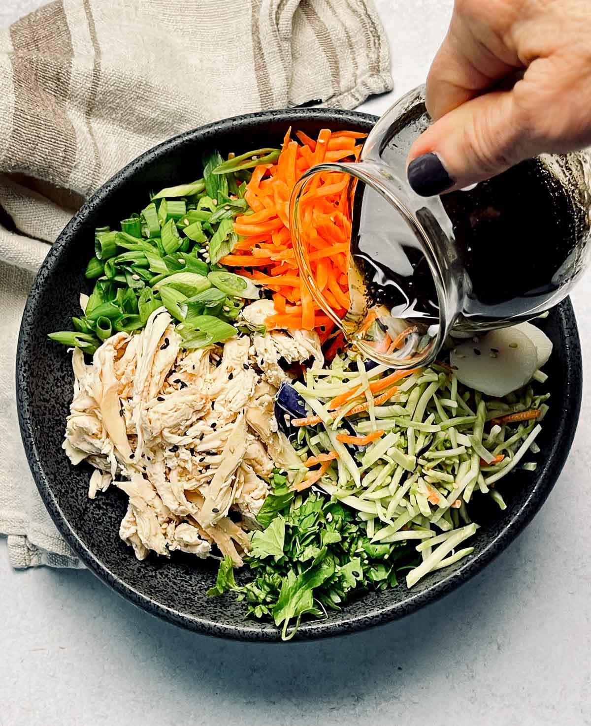 A hand pouring dressing into a large black bowl filled with Chinese chicken salad featuring shredded chicken, shredded carrots, broccoli slaw, scallions, water chestnuts and chow mein noodles, with a linen napkin on the side.
