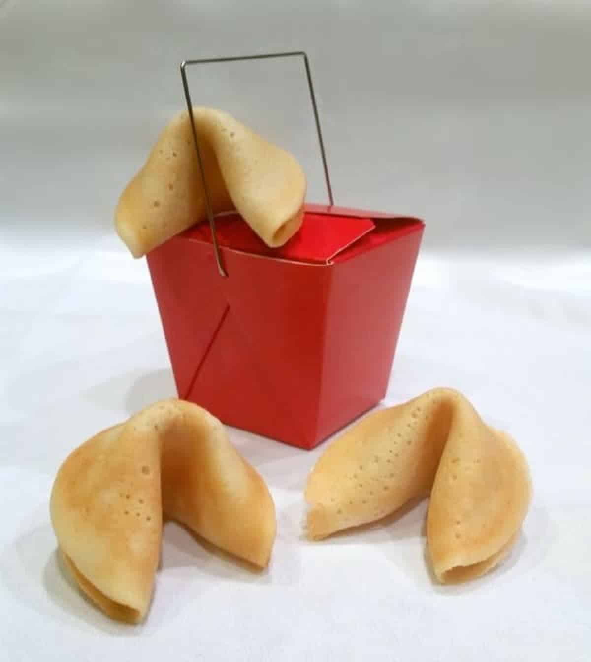 Homemade fortune cookies with a red Chinese takeout box on top of a white surface.