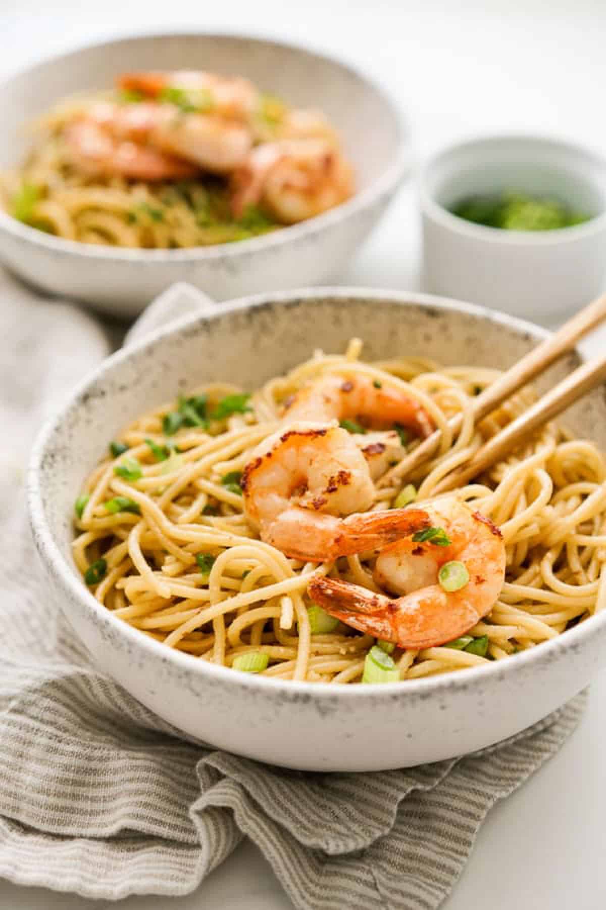 Garlic noodles with shrimp on top with chopsticks in a white bowl and another bowl in the background on top of a linen napkin.