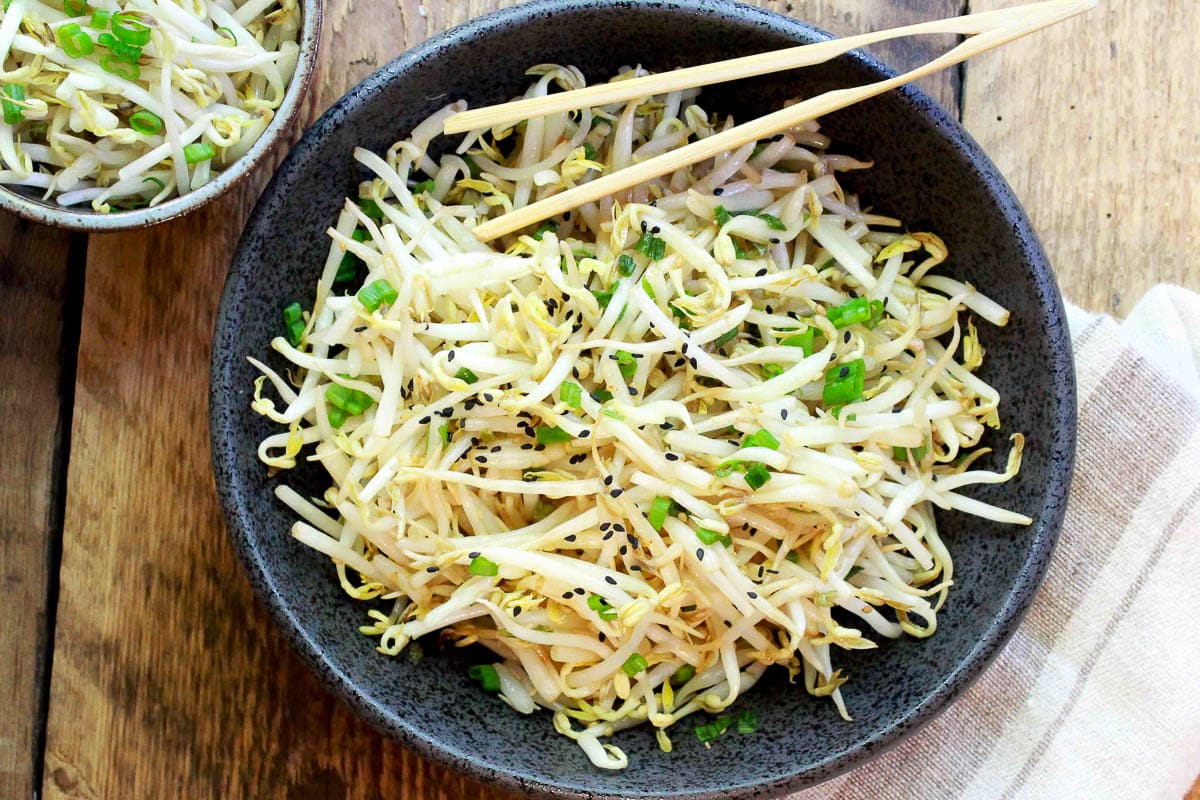 Japanese bean sprouts salad in a black bowl with chopsticks on top and a smaller bowl of bean sprouts on the side on top of a wooden board.