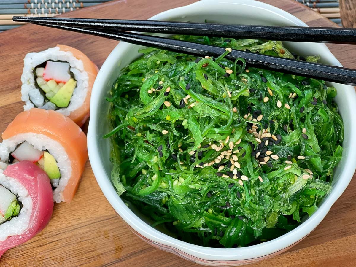 A bowl filled with Japanese seaweed salad with black chopsticks on top and assorted sushi on the side, on a wooden board.