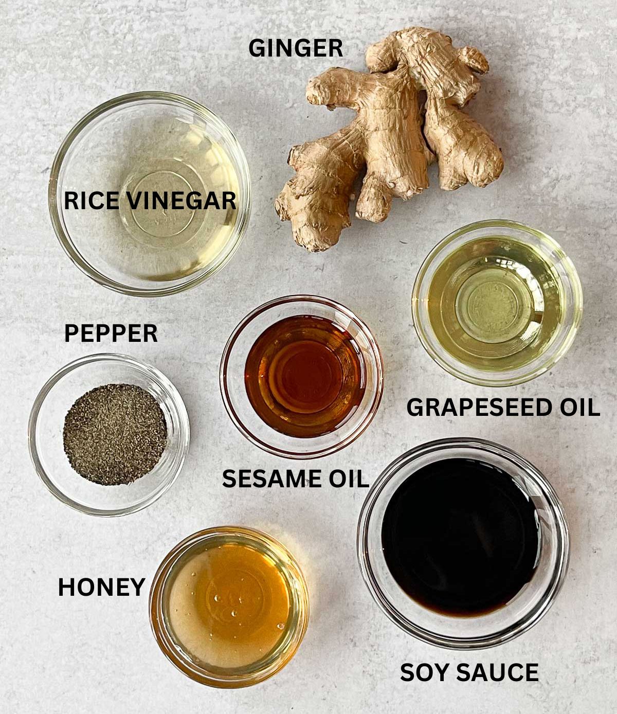 Labeled ingredients in small glass bowls for making the dressing for Chinese chicken salad along with a ginger root, on a gray surface.