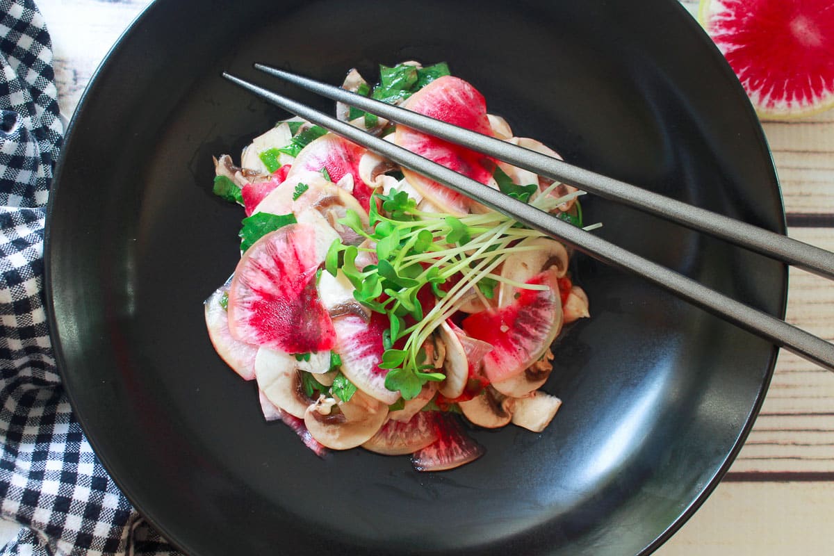 Mushoom and radish salad in a round black bowl with thinly sliced rainbow radishes, sliced mushrooms, and cilantro, topped with a yuzu vinaigrette, with a pair of silver chopsticks on top.