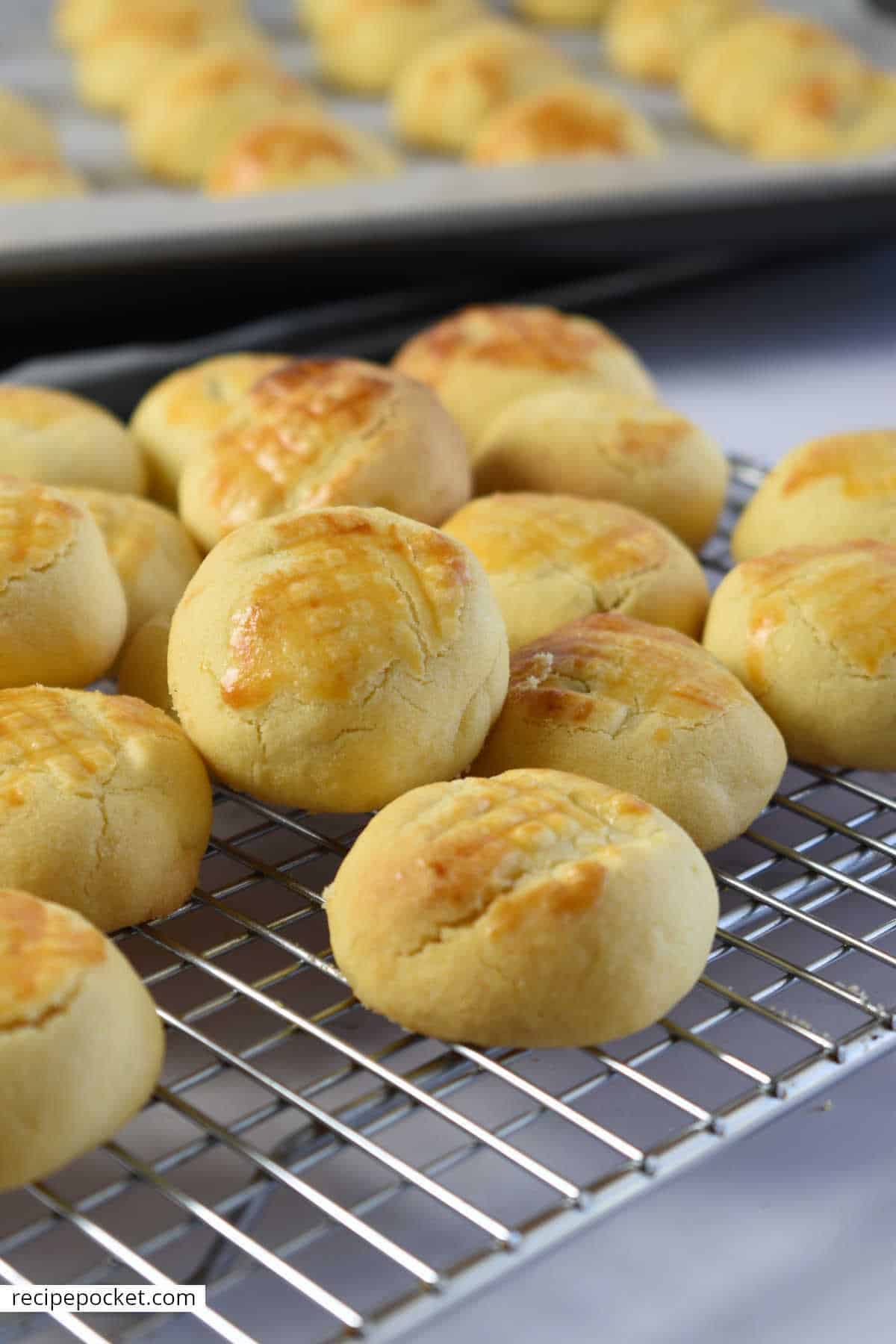 Golden Chinese pineapple tarts cooling on a baking rack with a batch of tarts on a baking tray in the background.