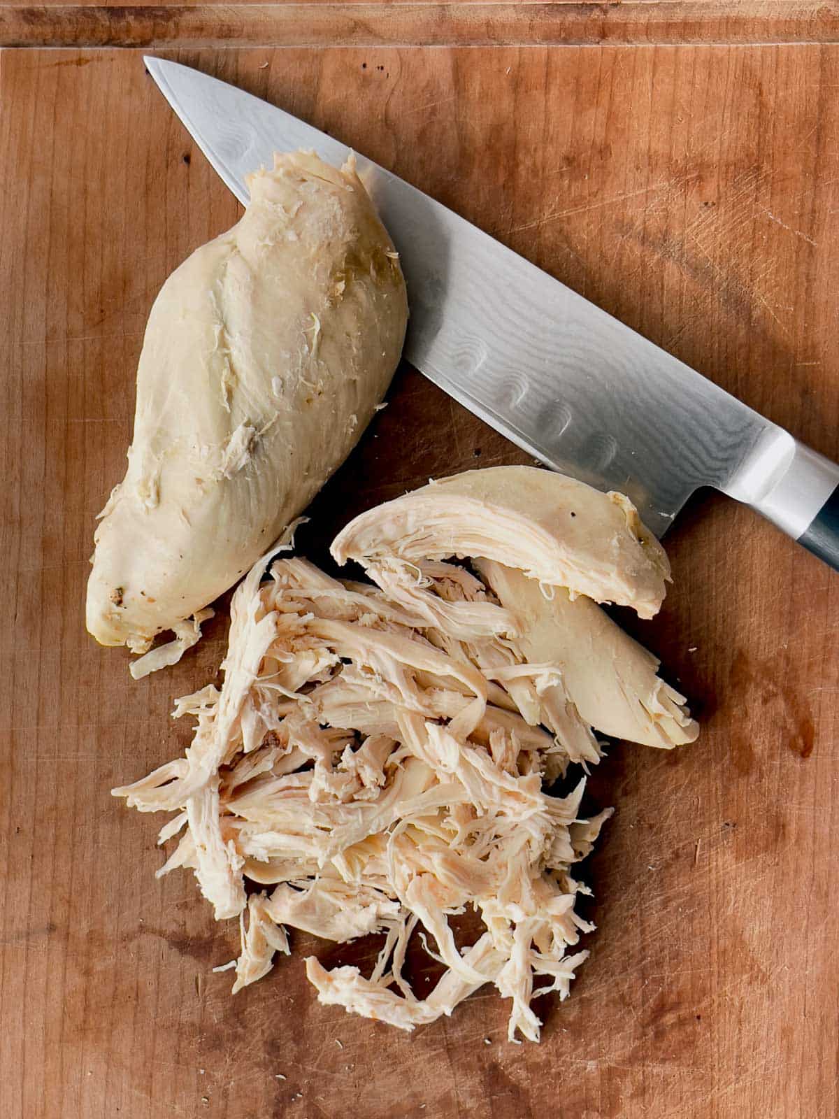A whole cooked chicken breast and a pile of shredded chicken breast on top of a wooden cutting board with a chef's knife on the side.