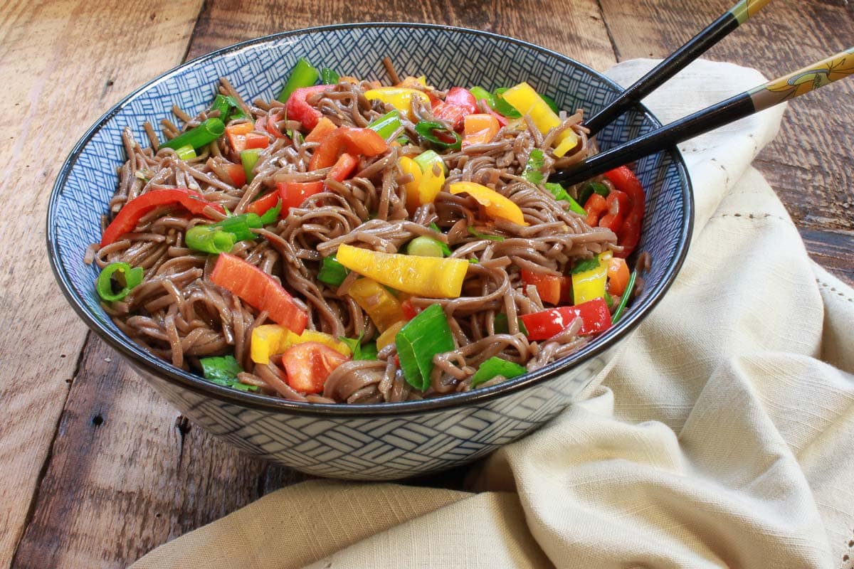 Colorful Japanese soba noodle salad in a large blue bowl with chopsticks on top of a wooden board and a napkin on the side