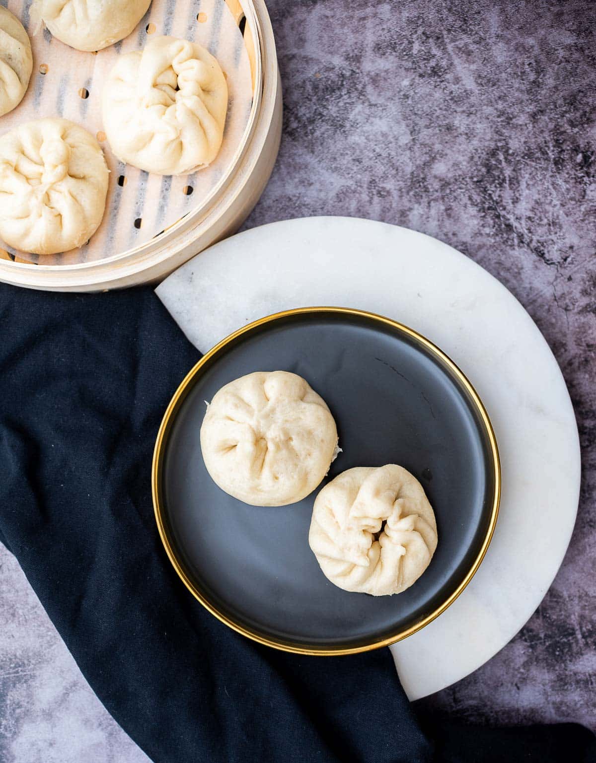 A round blue plate with two chinese steamed pork buns and a bamboo steamer on the side placed on top of a gray surface.