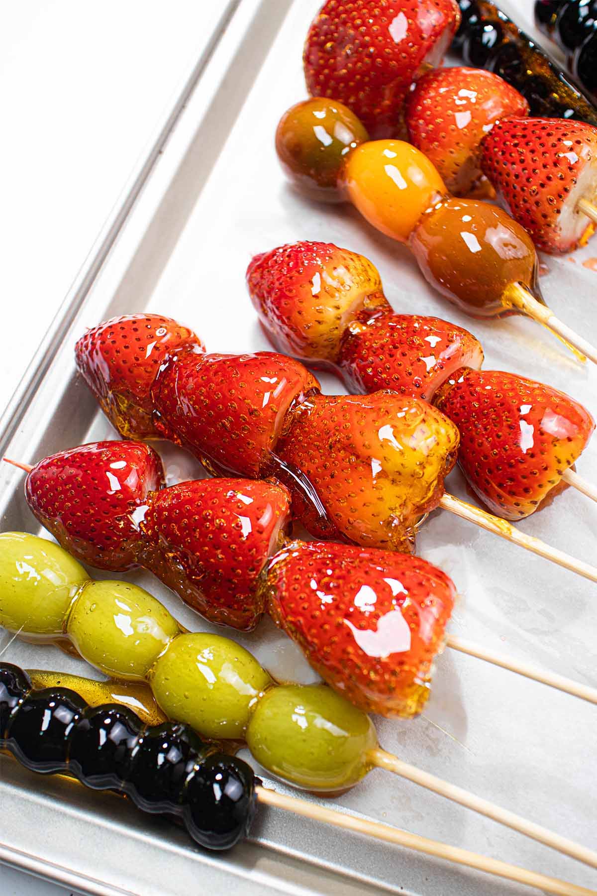 Skewers of Tanghulu Chinese candied fruit on a baking sheet including skewers of strawberries, grapes, and blueberries.