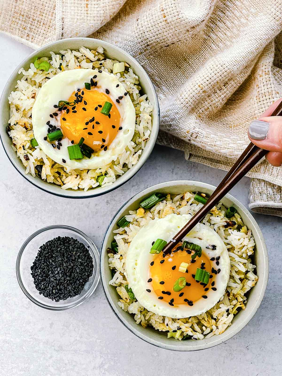 Crispy rice with ginger and scallions topped with a runny fried egg in two small bowls with a pair of brown chopsticks, a linen napkin, and black sesame seeds in a bowl on the side.