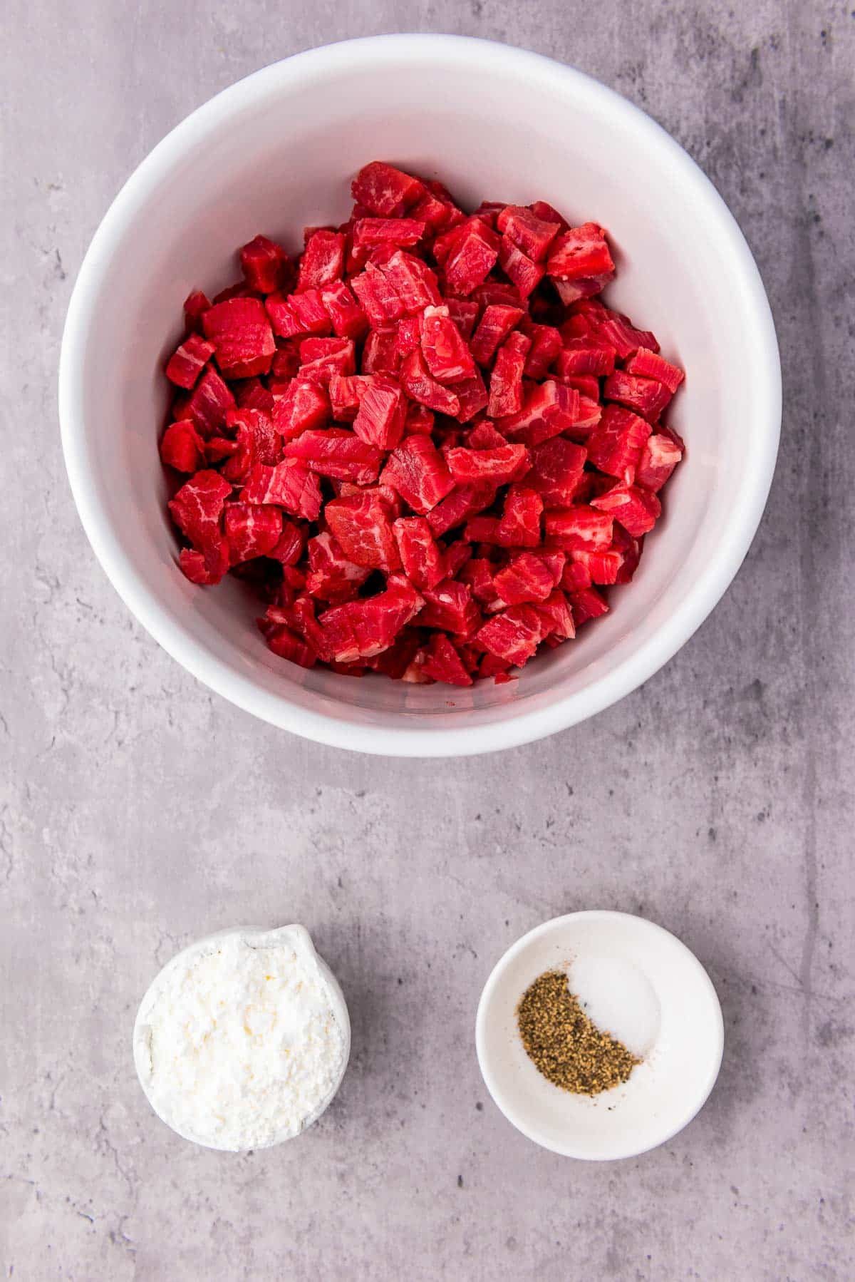 Cubed flank steak in a white bowl with two white bowls of salt and pepper on the side.