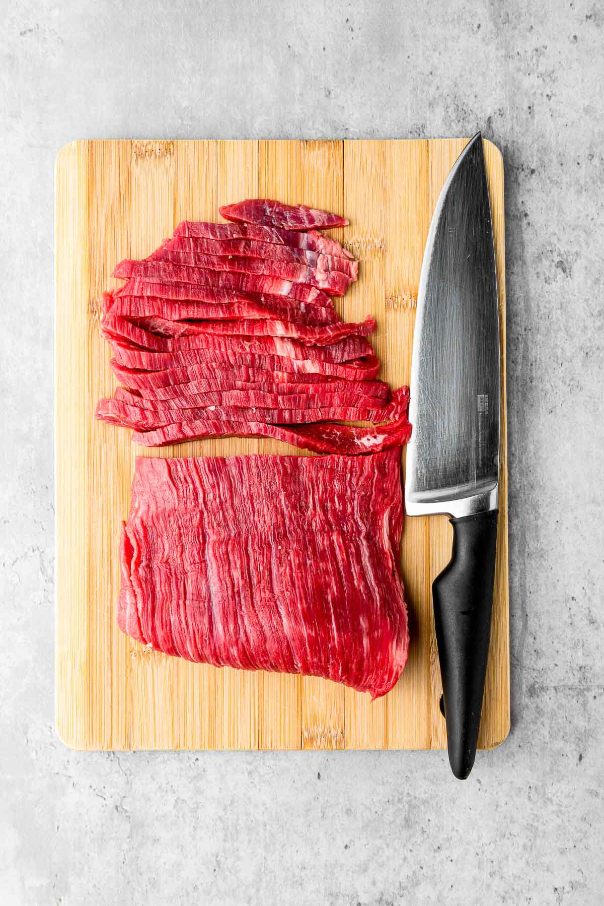 Sliced flank steak on top of a wooden cutting board with half an uncut steak and a large knife on the side.