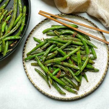 Chinese Garlic Green Beans on a round white plate with a pair of brown chopsticks and green beans in a frying pan on the side with a napkin.