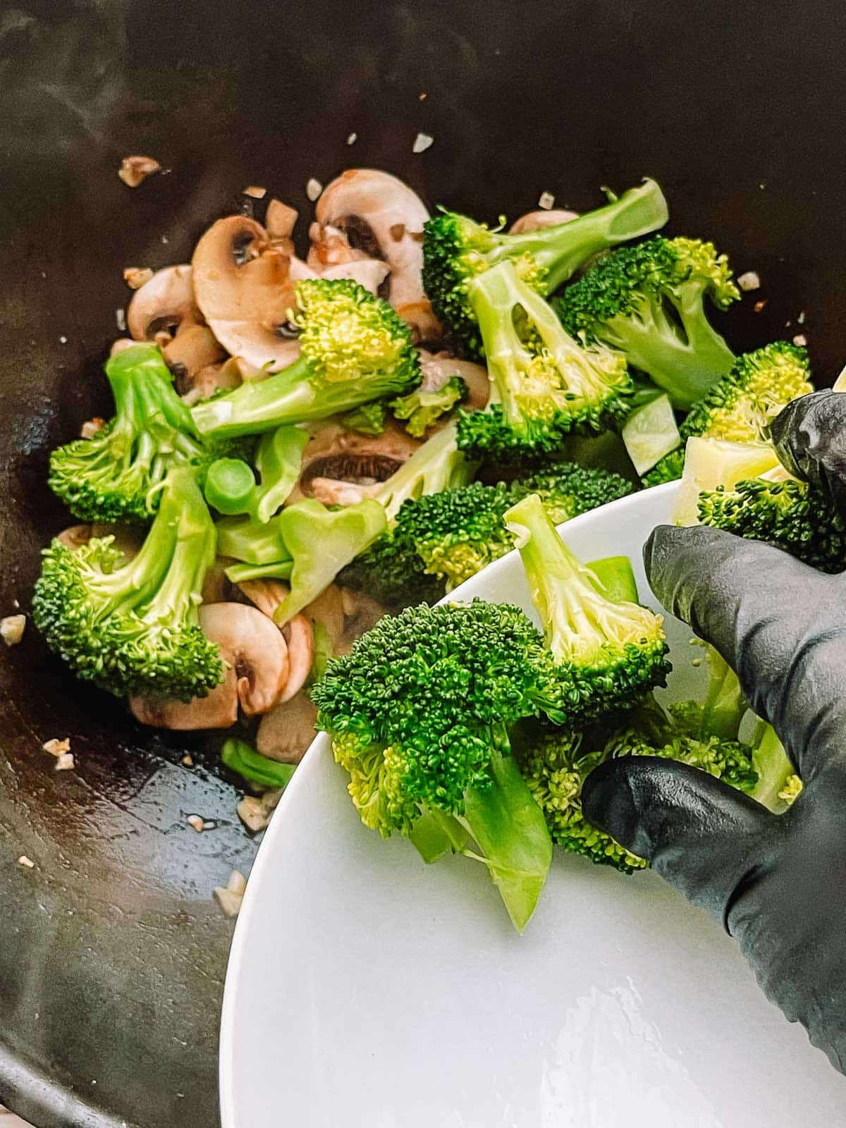 Adding broccoli florets to a wok with stir fried button mushroom slices.