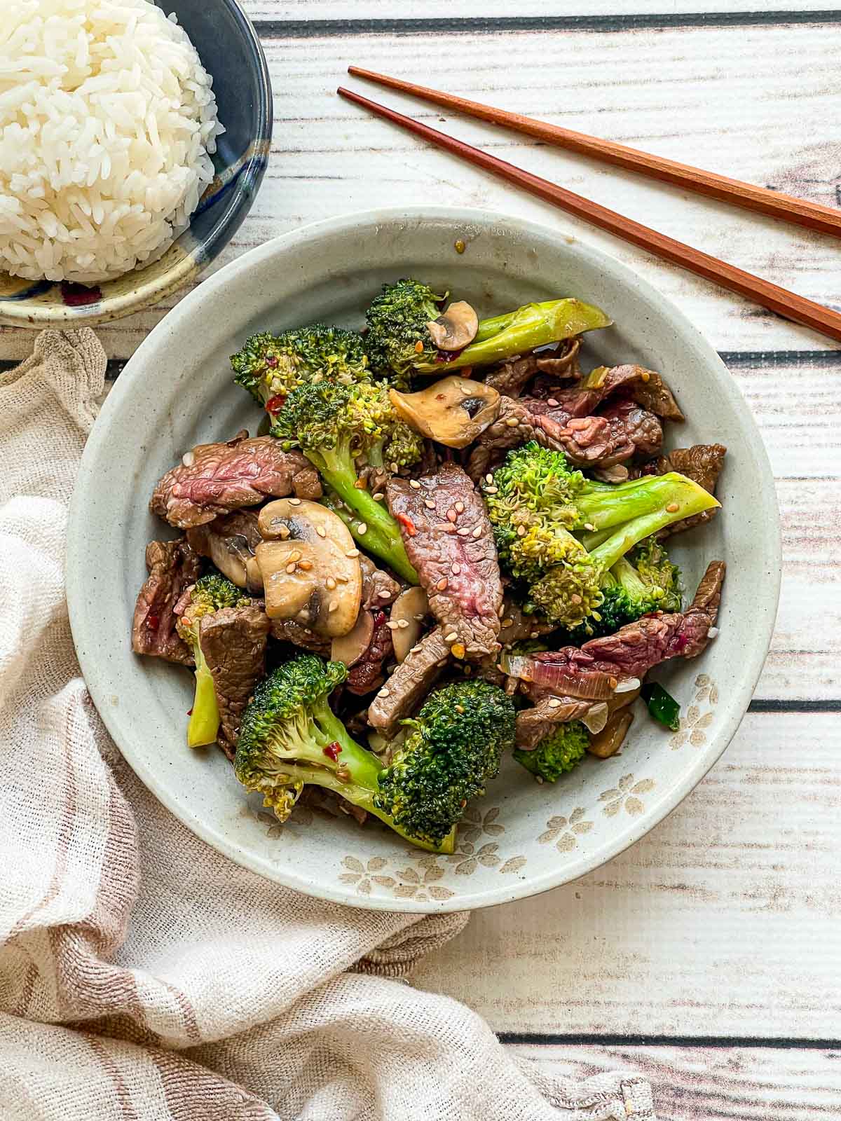 Beef and broccoli stir fry in a round cream-colored bowl with a pair of wooden chopsticks and a small bowl of white rice on the side on top of a white wooden board.