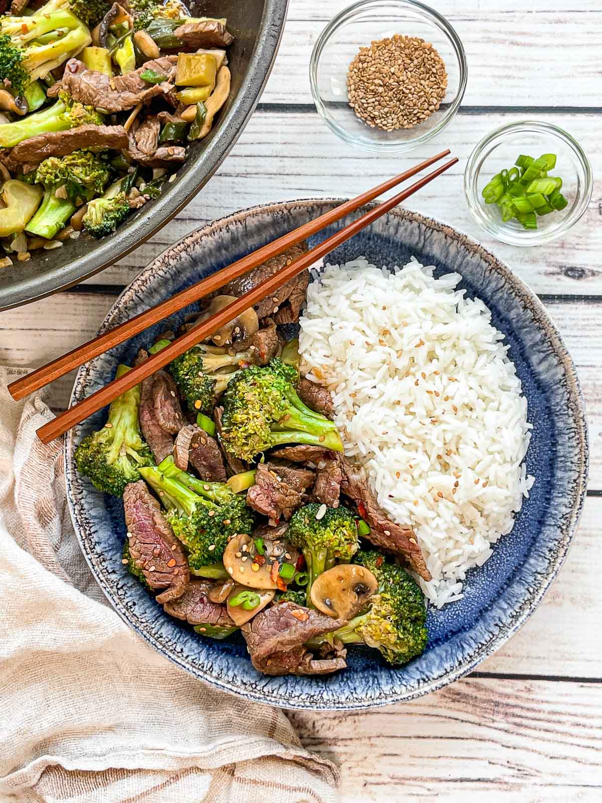 Beef and broccoli stir fry and white rice in a round blue bowl with a pair of wooden chopsticks on top and a wok filled with stir fry, and two small glass bowls with sesame seeds, and sliced green onions, and a linen napkin on the side, on top of a white wooden board.