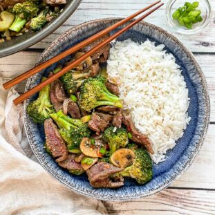 Beef and broccoli stir fry and white rice in a round blue bowl with a pair of wooden chopsticks on top and a wok filled with stir fry, and a small glass bowl with sliced green onions, and a linen napkin on the side, on top of a white wooden board.