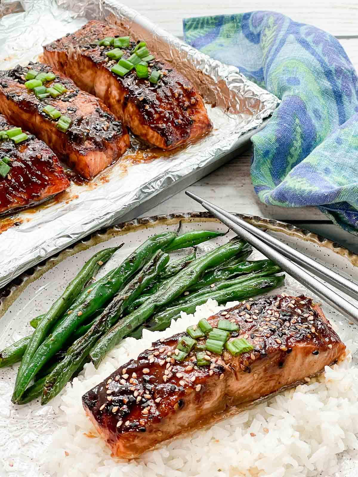 A filet of broiled miso glazed salmon on top of white rice on a plate with silver chopsticks and blistered green beans and a baking tray with broiled miso glazed salmon filets and a blue napkin on the side.