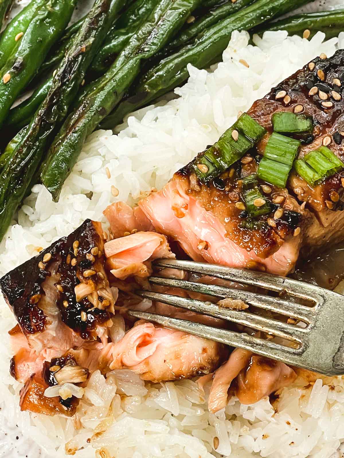 A fork digging into a piece of broiled miso glazed salmon on top of white rice on a plate with a side of blistered green beans.