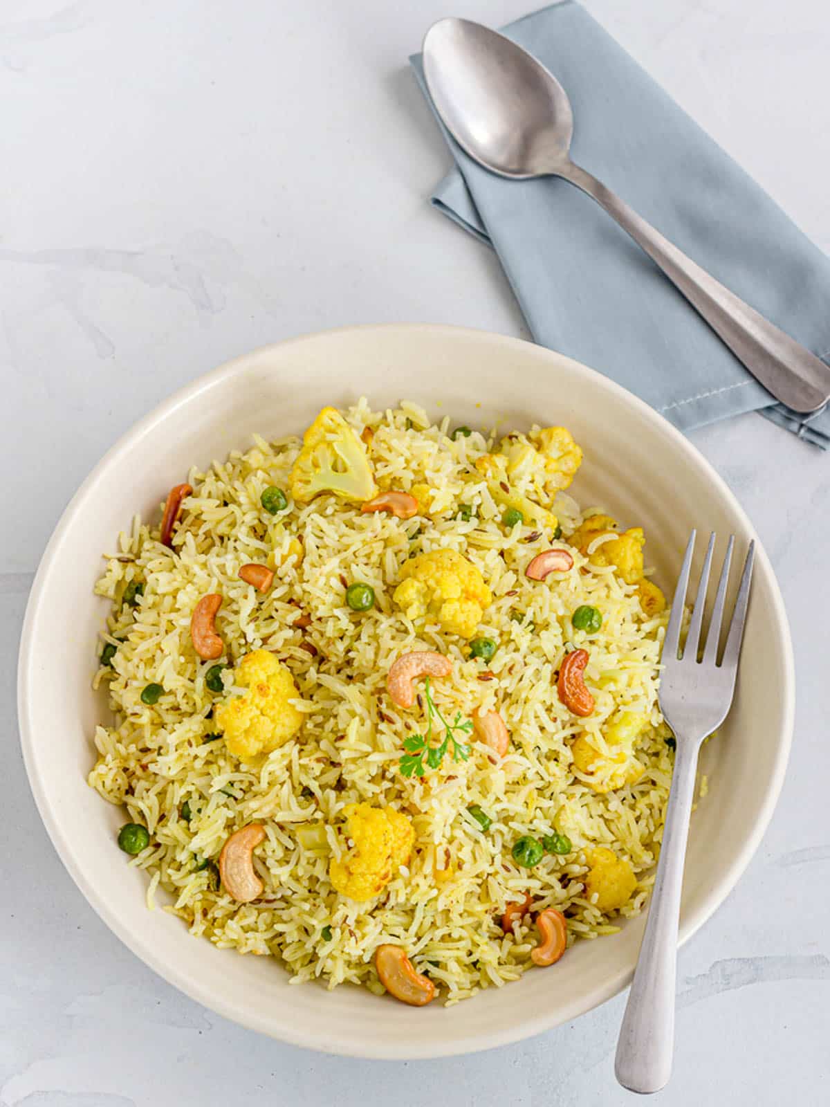 Air fryer veggie fried rice in a round white bowl with a fork, and a spoon on top of a blue napkin on the side, on a gray surface.