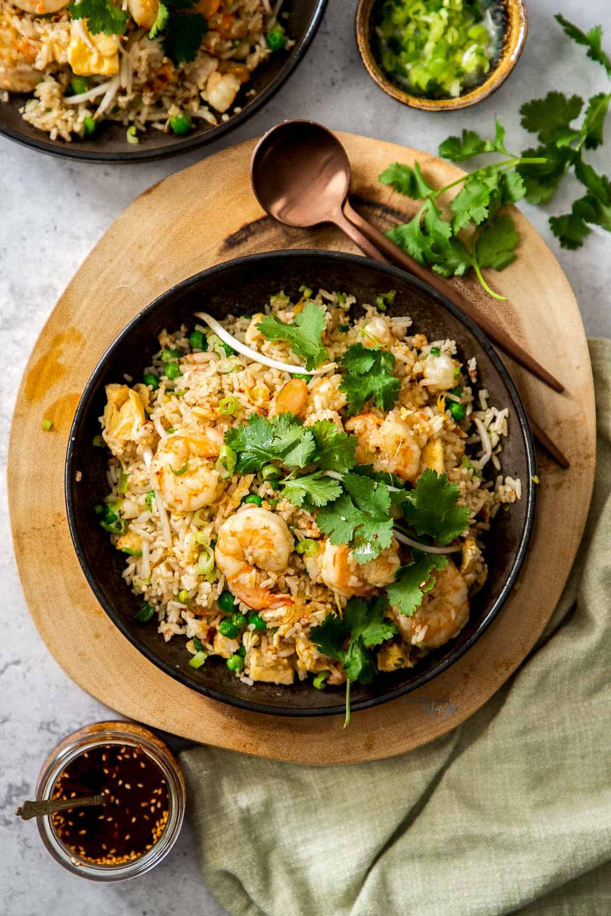 Chinese Prawn Fried Rice topped with cilantro leaves in a round black bowl on top of a wooden board with a copper spoon on the side, and a side of sauce and toppings and a linen napkin in small bowls on top of a gray surface.