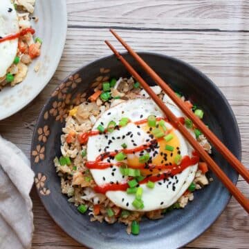 Cauliflower fried rice with crab topped with a runny egg in a round dark gray bowl with wooden chopsticks on top on a white wooden board.