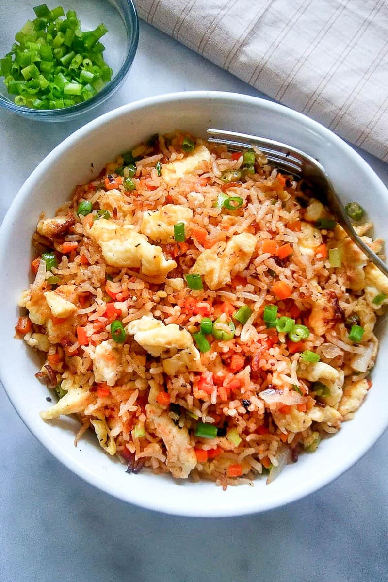 Egg fried rice in a round white bowl with a fork, and a small glass bowl of sliced green onions on top of a gray surface with a linen napkin on the side.