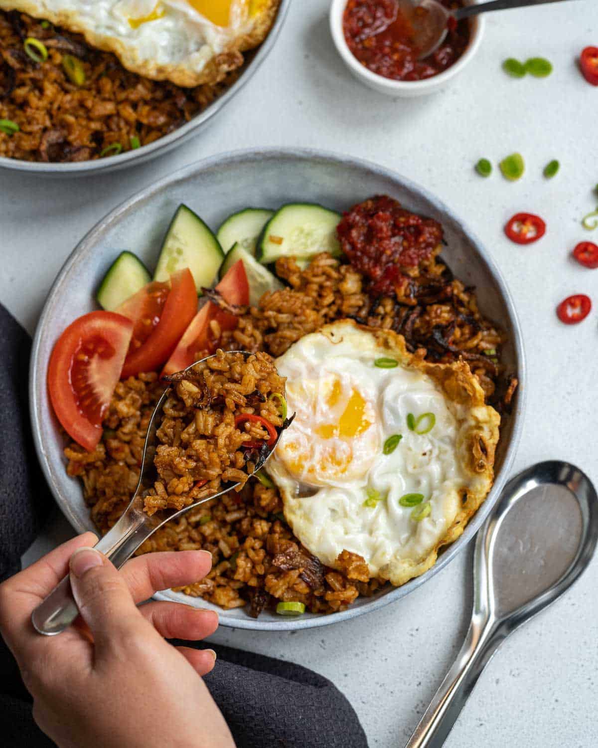 A hand with spoonful of Indonesian fried rice (nasi goreng) above a round gray bowl of fried rice topped with a runny egg and a side of sliced tomatoes and cucunmber, on a gray surface.