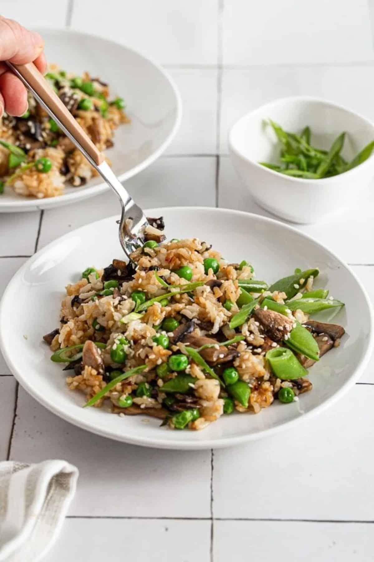 A hand lifting a forkful of mushroom fried rice in a round white bowl with a small bowl of peas on the side on top of a white tile surface.