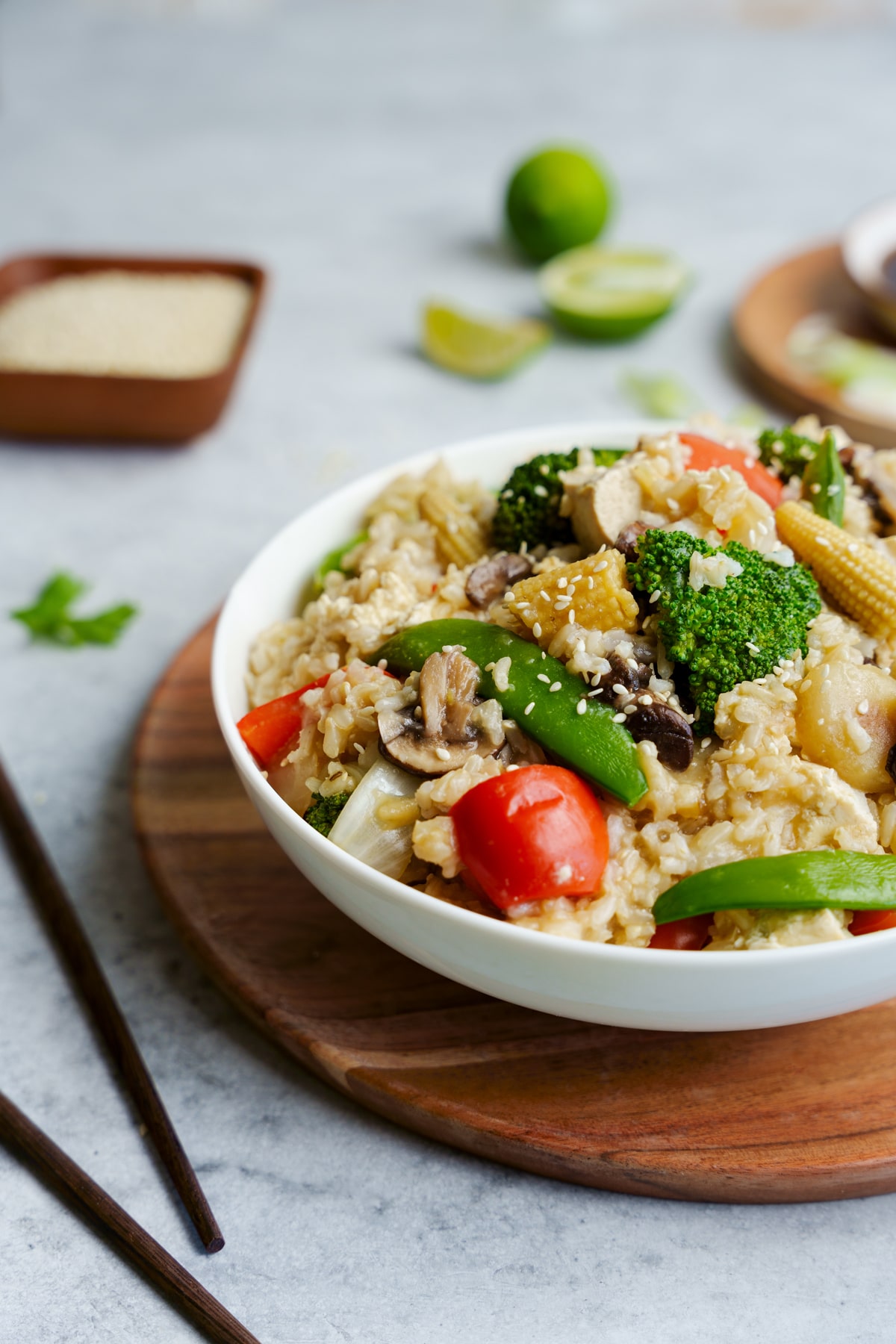 Tofu vegetable fried rice in a round white bowl on top of a round wooden board with brown chopsticks, sauce, and lime wedges on the side, on top of a gray surface.