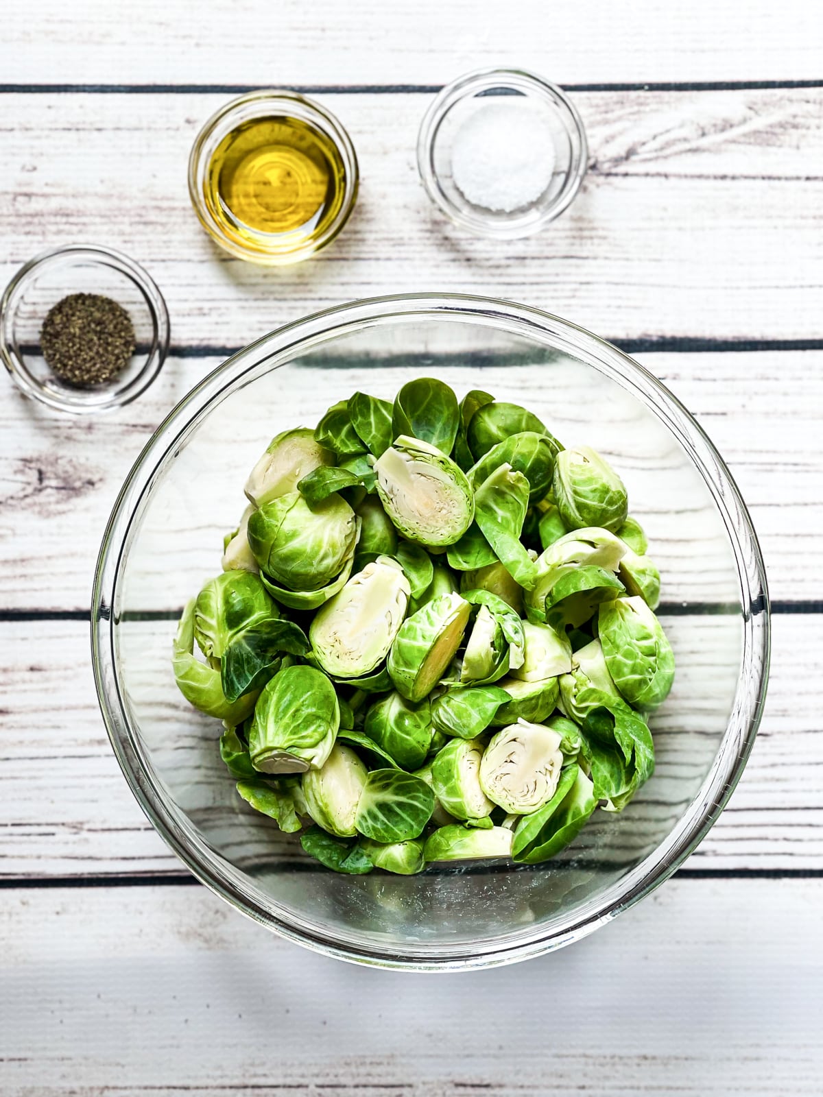Sliced, raw brussels sprouts in a large glass bowl with pepper, olive oil, and salt in small glass bowls on the side on top of a white board.