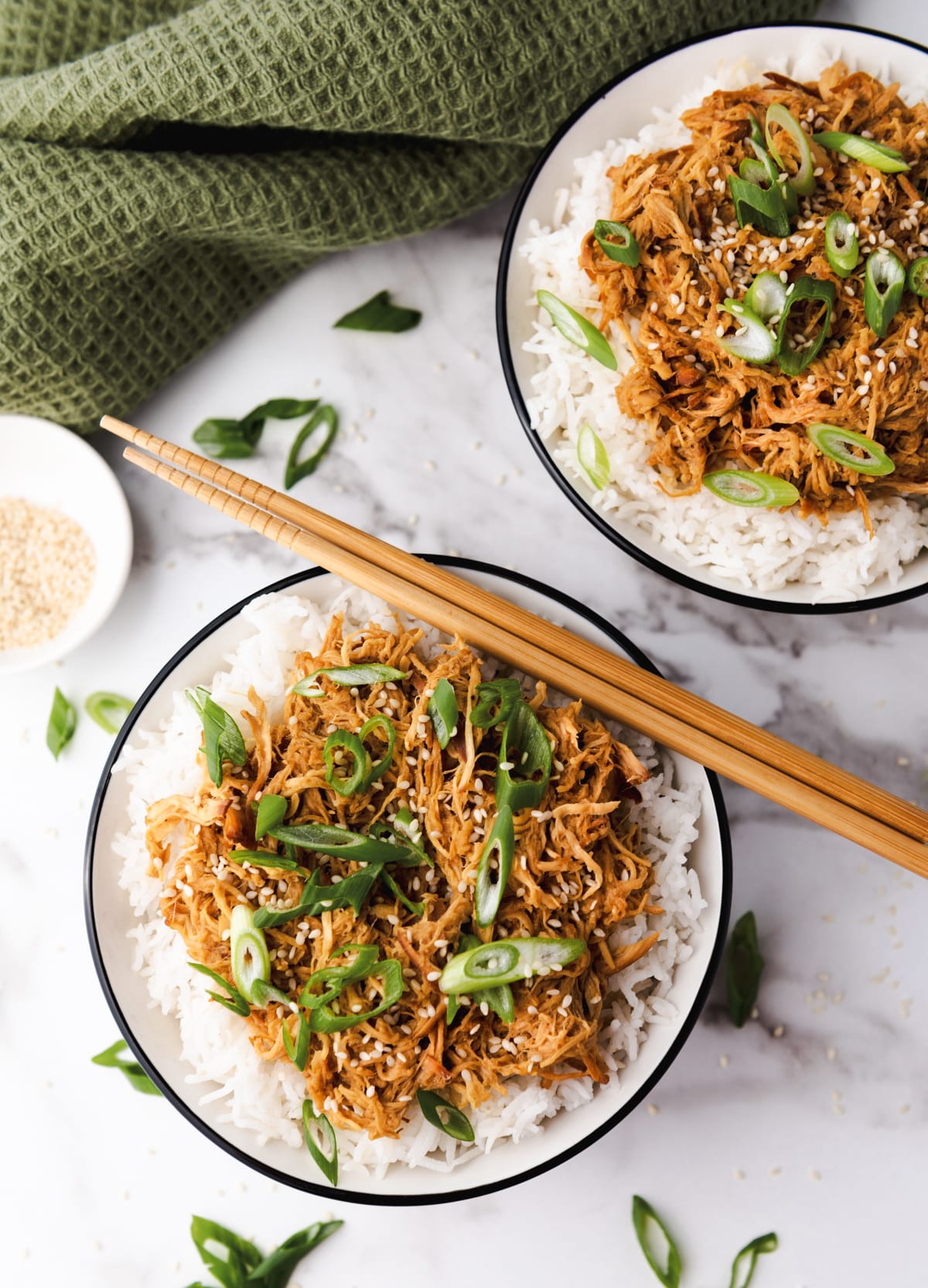 Slow cooker honey garlic chicken over white rice topped with sliced scallions placed in two white bowls with a pair of chopsticks on top and a small bowl of sesame seeds and a green napkin on the side, on top of a marble surface.