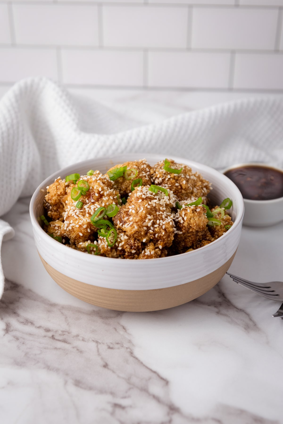 Air Fryer Teriyaki Cauliflower Wings topped with sesame seeds and sliced scallions in a white bowl with a side of teriyaki sauce in a small white bowl on top of a marble surface with a white napkin.