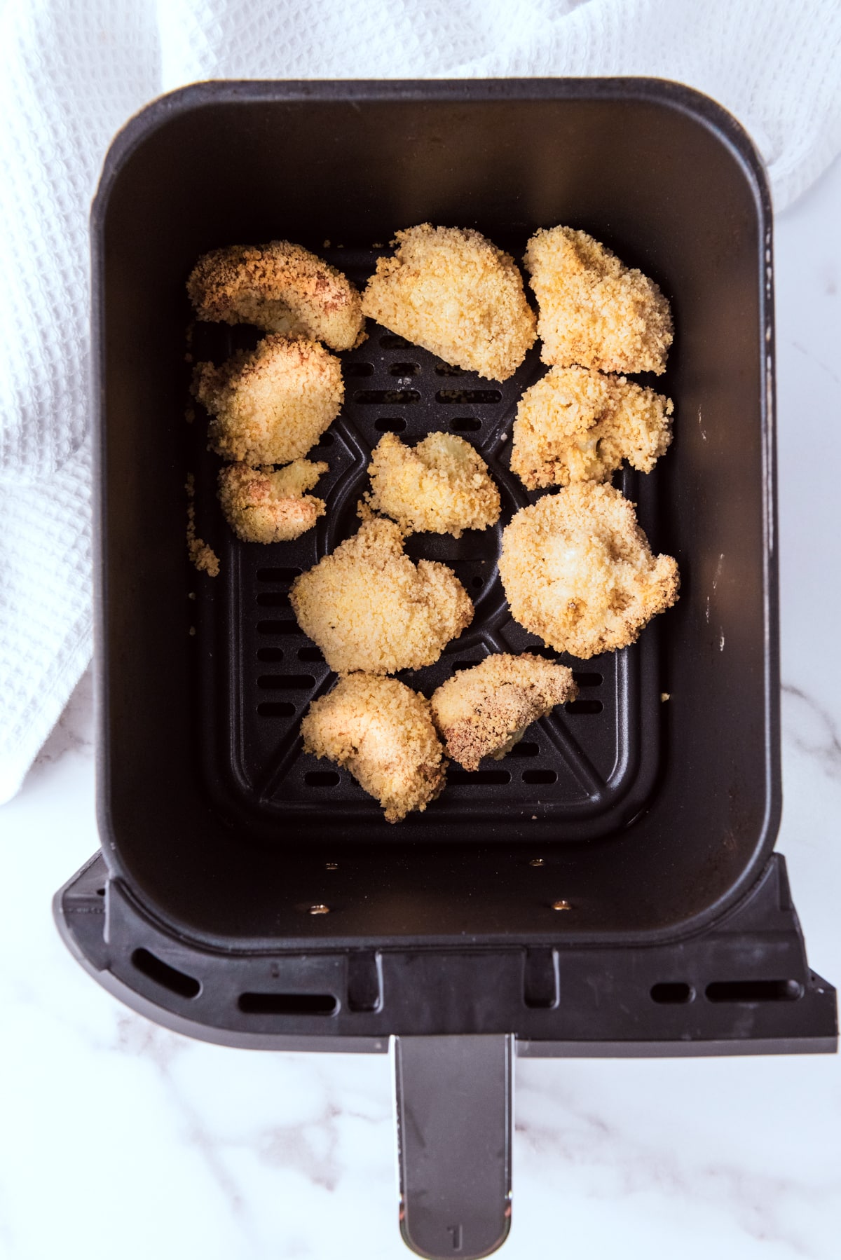 Breaded cauliflower florets in an air fryer basket on top of a marble surface with a white napkin.