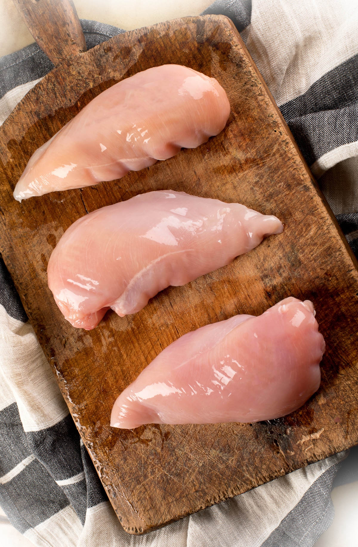 Three raw chicken breasts on a wooden cutting board on top of a black and white napkin.