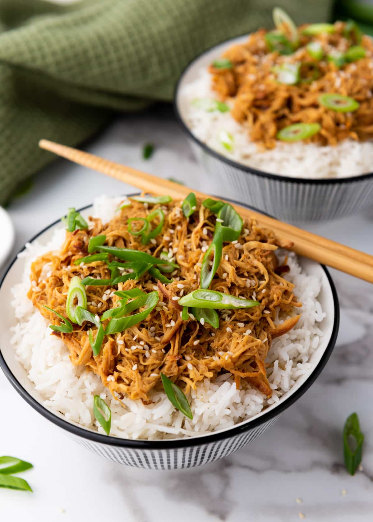 Slow cooker honey garlic chicken over white rice topped with sliced scallions placed in two white bowls with a pair of chopsticks on top and a green napkin on the side, on top of a marble surface.