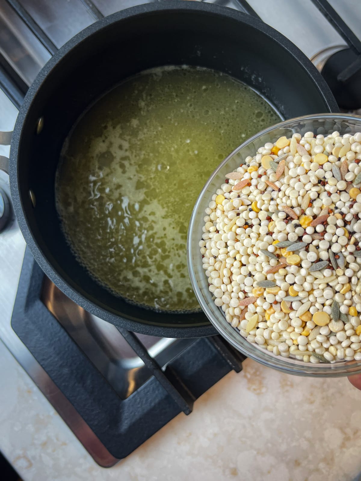 Couscous quinoa blend being put into a pot of chicken broth on a stove.