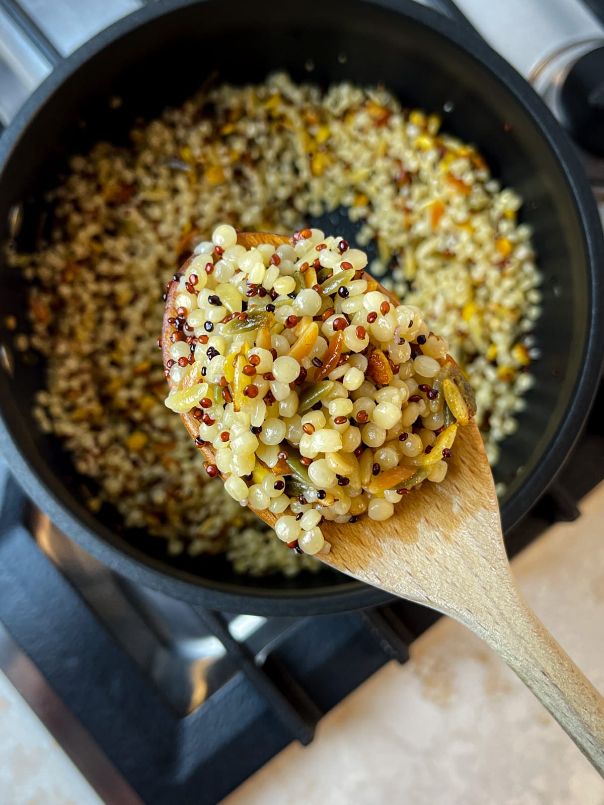 A wooden spoon holding up a blend of couscous and quinoa over a pot on a stove.