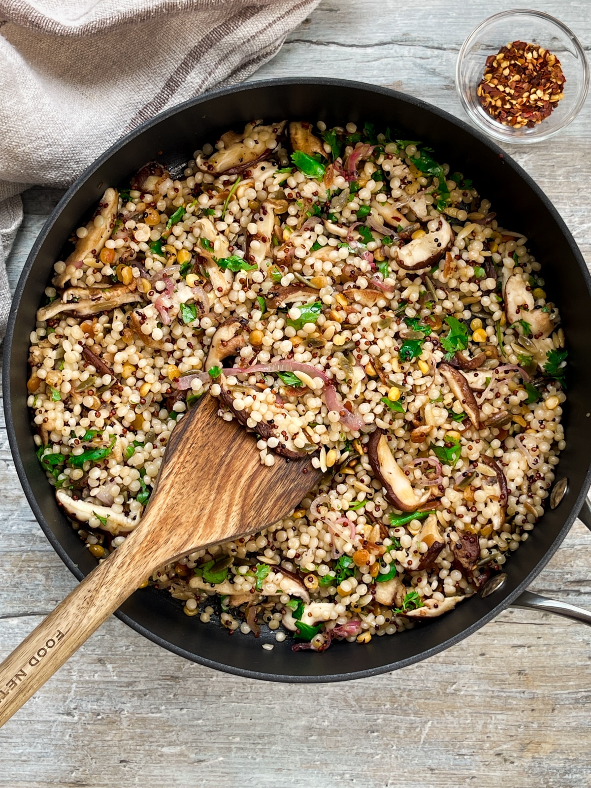 A wooden spatula inserted into a frying pan filled couscous and quinoa with shiitake mushrooms on top of a wooden board with a linen napkin and red pepper flakes on the side.