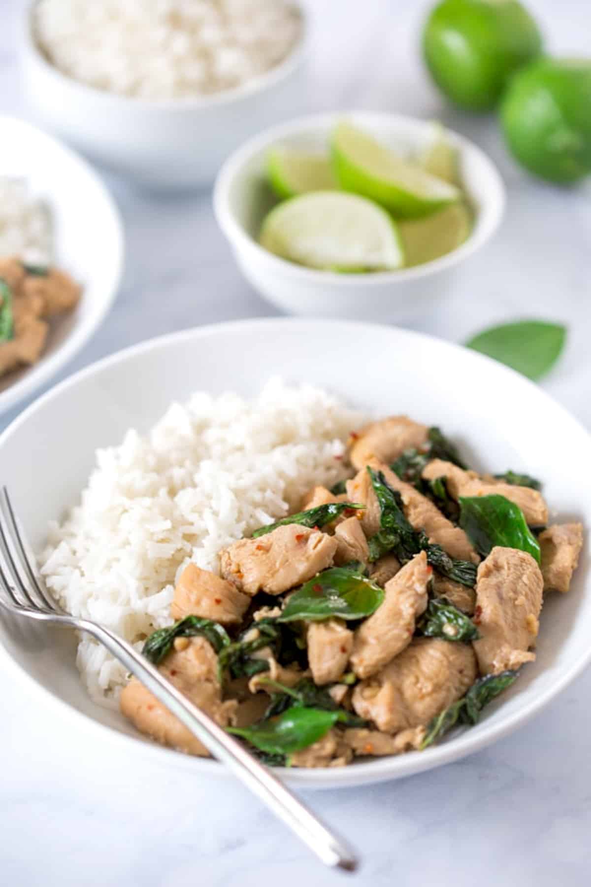Thai basil chicken in a white bowl with white rice and basil on top, and a bowl of lime wedges, a bowl of white rice, and whole limes on the side.