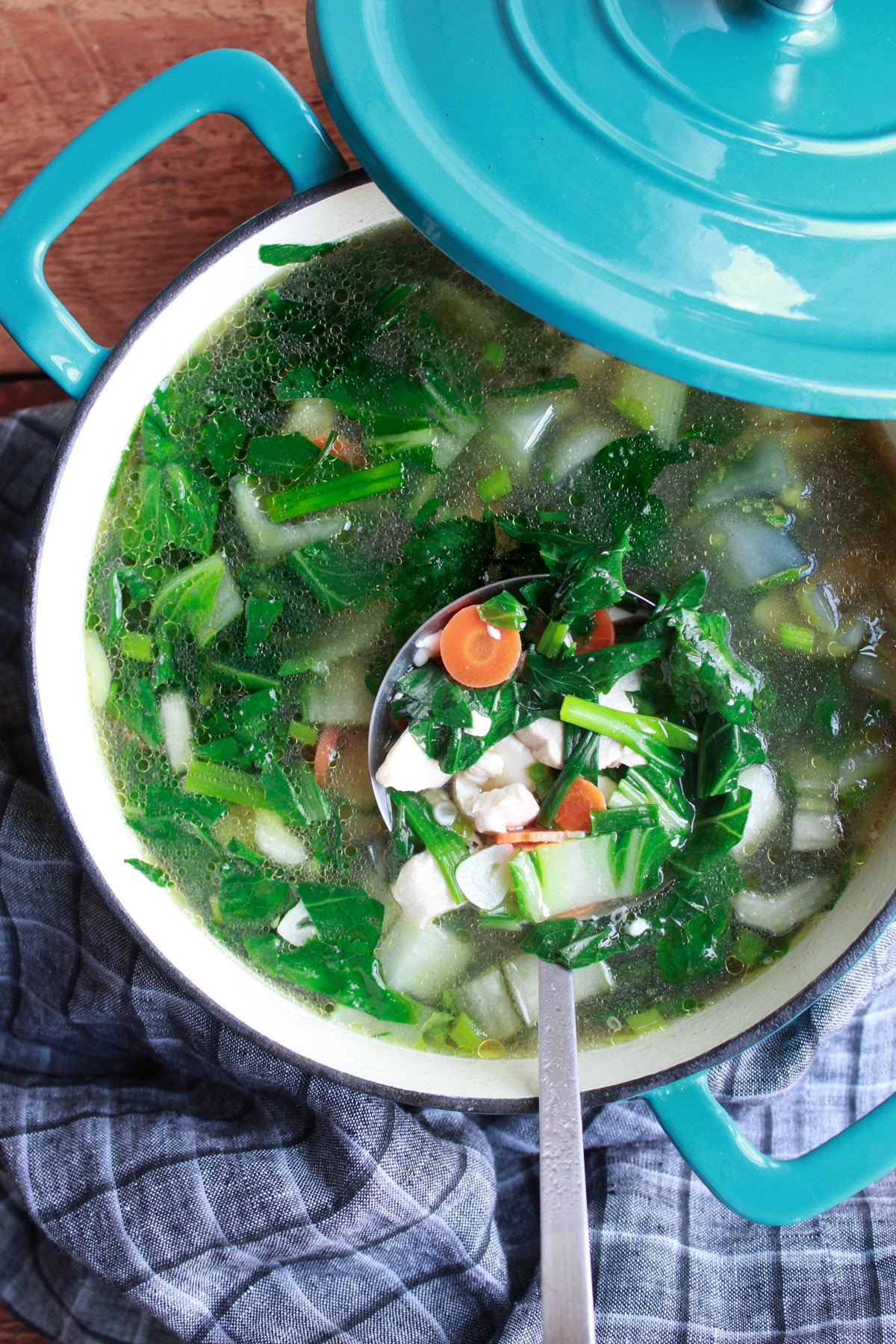 Thai clear soup with chicken and tofu in a large blue soup pot with a ladle inserted and a lid and gray napkin on the side, on top of a wooden board.