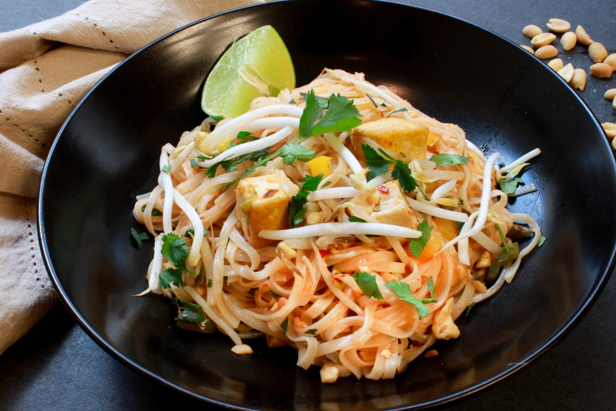 Tofu Pad Thai presented in a black bowl topped with bean sprouts with a side of lime wedge on top of a black surface with a linen napkin and peanuts on the side.