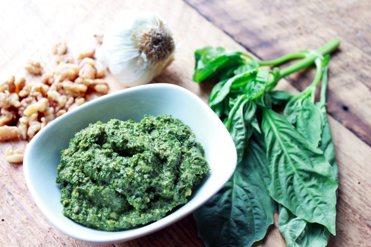 Thai Basil Mint Cilantro Pesto in a small white bowl on top of a wooden board with fresh basil leaves, a garlic bulb, and raw walnut pieces on the side.