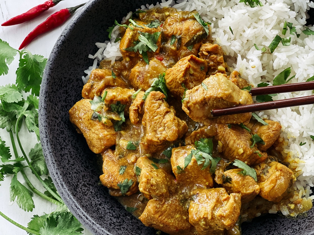 A black bowl of Thai curry chicken with white rice and chopsticks with fresh cilantro and red chili peppers on the side, on top of a white board.