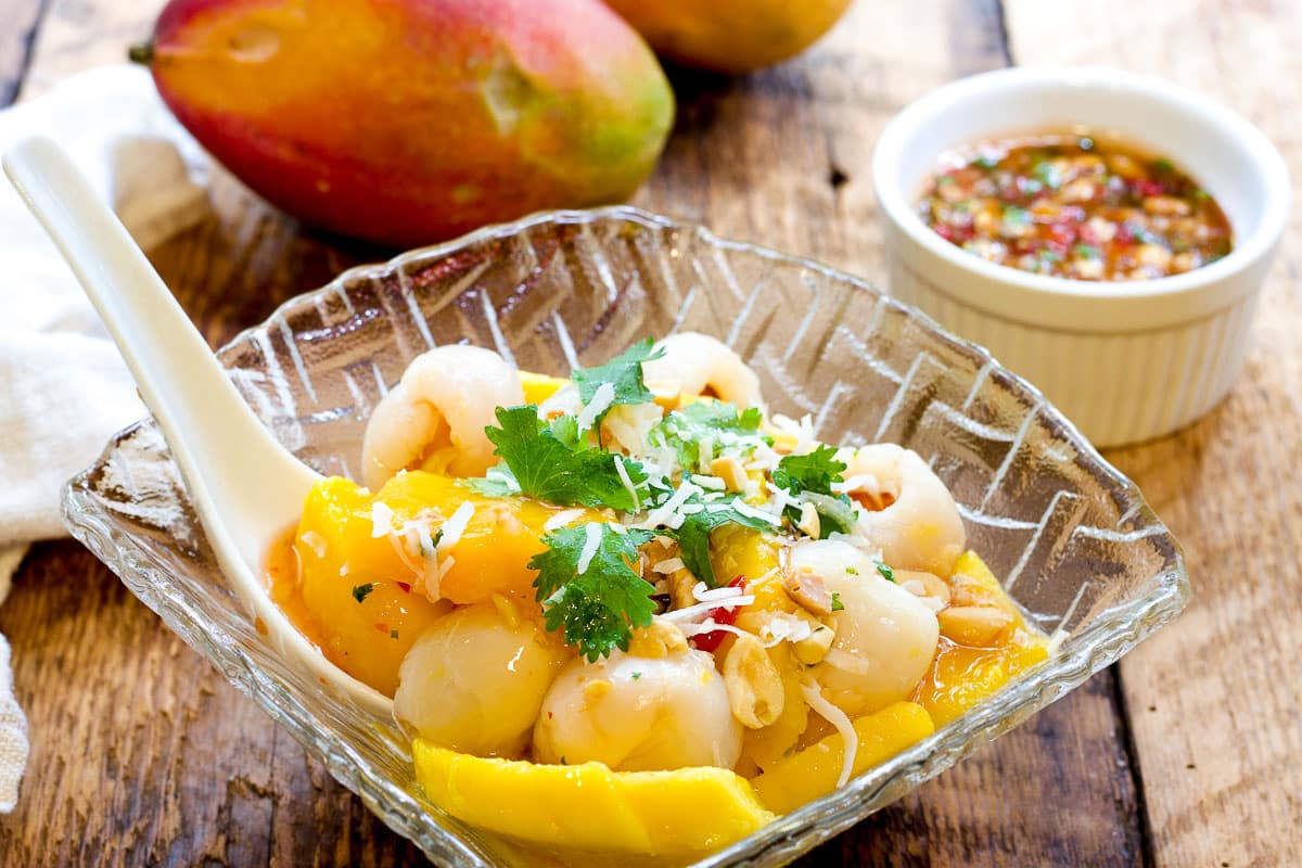 Thai Mango and Lychee Salad in a glass bowl topped with toasted coconut flakes and fresh cilantro, on top of a wooden board with a small white bowl of sweet and spicy sauce and whole mangos on the side.
