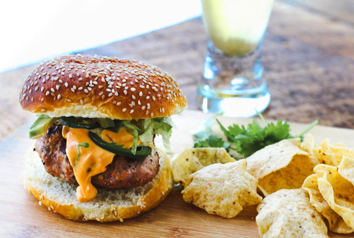 A Thai Turkey Burger on a sesame bun topped with a spicy aioli and shredded lettuce, with tortilla chips on the side and a glass of beer in tje background.