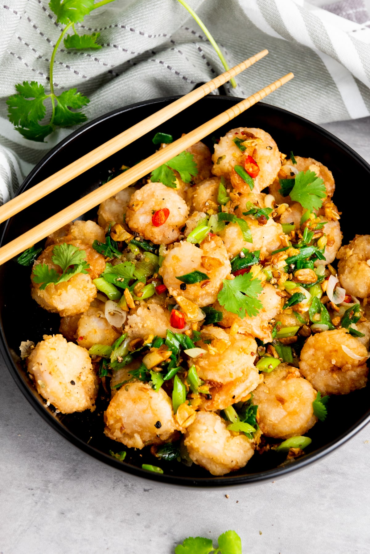 Chinese salt and pepper shrimp in a black bowl with sautéed vegetables and herbs, and a pair of chopsticks on top on a gray surface with a gray linen on the side.