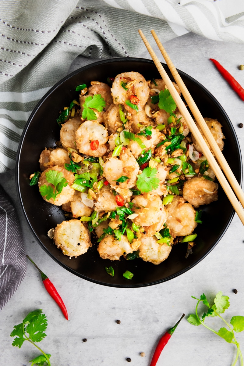 Chinese salt and pepper shrimp in a black bowl with sautéed vegetables and herbs, and a pair of chopsticks on top on a gray surface with a gray linen on the side.