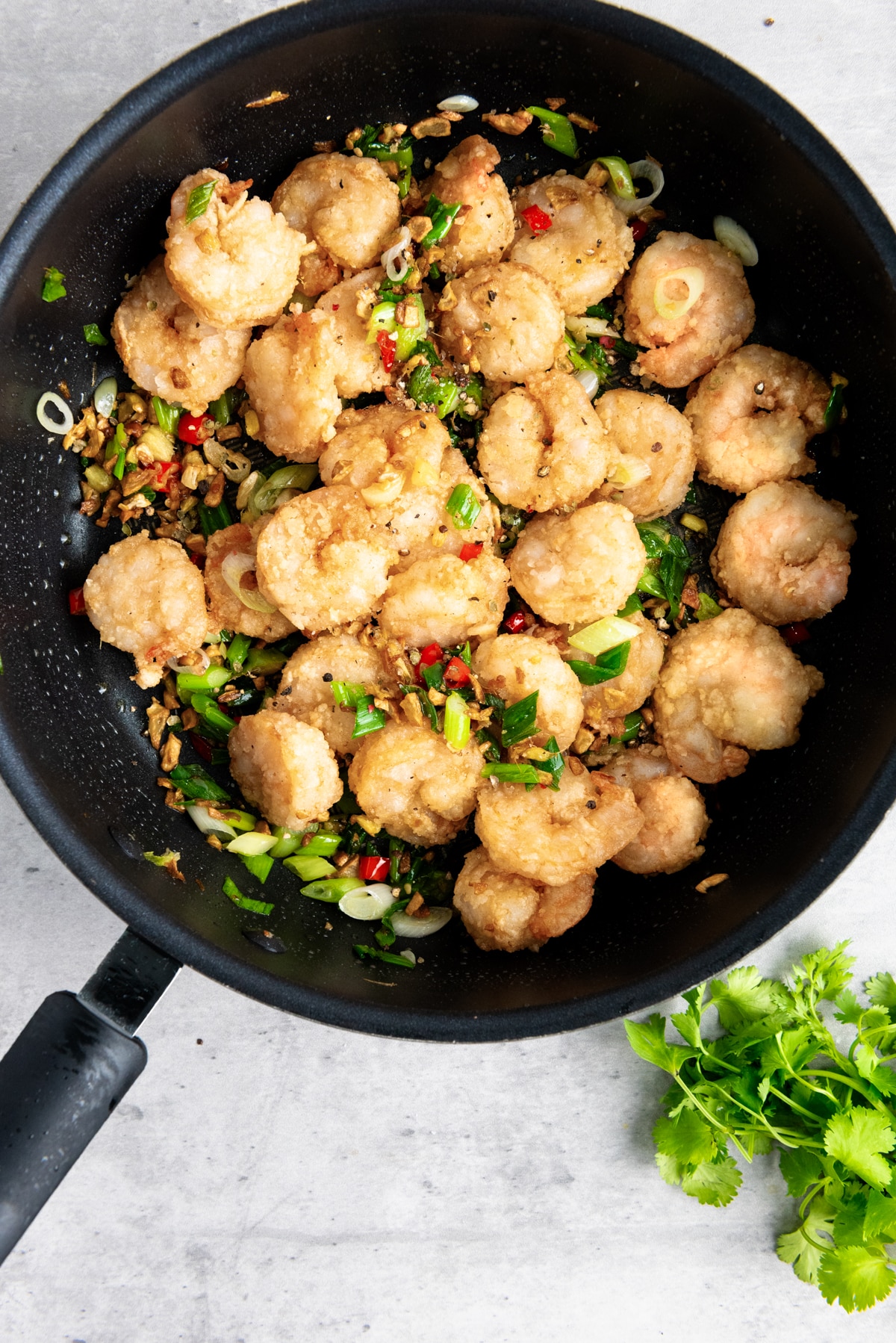 Chinese salt and pepper shrimp in a frying pan with sautéed vegetables and herbs on top on a gray surface with fresh cilantro on the side.