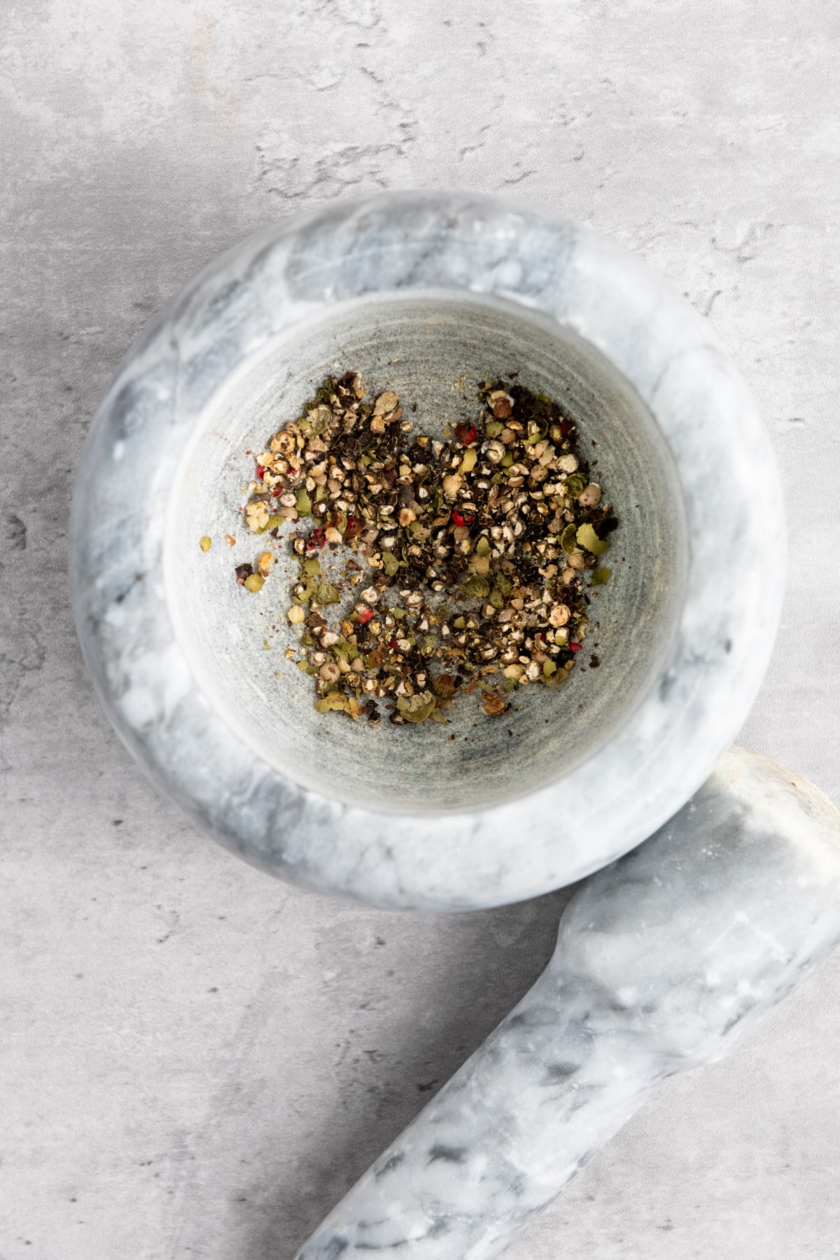 Crushed whole peppercorns inside a gray ceramic mortar bowl with a ceramic pestle on the side on top of a gray surface.
