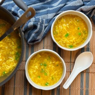 Chinese egg drop soup in two bowls and a pot with a white spoon and blue napkin on the side on top of a bamboo mat.